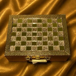 May include: A gold-colored, rectangular box with a checkerboard pattern on the lid. The checkerboard squares alternate between a reflective, mirrored surface and a textured, glittery gold. The box has a gold clasp and hinges.