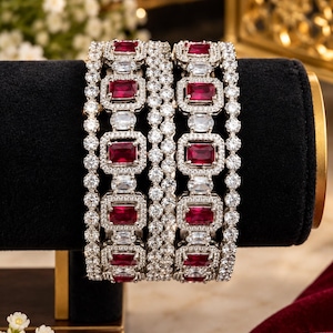 Op de afbeelding: Twee zilverkleurige armbanden met rechthoekige donkerrode stenen in een zetting van kleine heldere kristallen. Het ontwerp bevat afwisselende rijen van heldere ovale stenen en pavé-gezette kristallen, wat zorgt voor een gestructureerd uiterlijk.