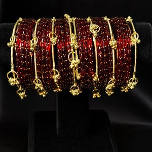 Pode incluir: Um conjunto de pulseiras de vidro vermelho com detalhes dourados e pingentes pendurados. As pulseiras são exibidas em um suporte preto sobre um fundo preto, destacando seu design detalhado e cor rica.