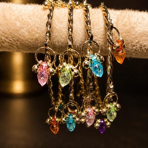 Puede incluir: Pulseras doradas con colgantes de cristal facetado de colores. Cada pulsera presenta un diseño de cadena trenzada y pequeñas cuentas doradas redondas. Los colgantes son en tonos rosa, verde, azul, naranja y morado.