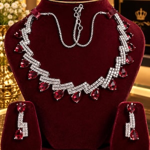 May include: A silver-toned necklace and earring set featuring a zigzag pattern encrusted with clear crystals and accented by dangling, pear-shaped deep red stones. The necklace includes a delicate chain with an adjustable clasp for length.
