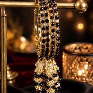 May include: A set of three gold-toned bangles featuring alternating black and clear faceted beads. Each bangle is adorned with a dangling cluster of matching black and clear beads at the bottom, creating a layered and textured appearance.