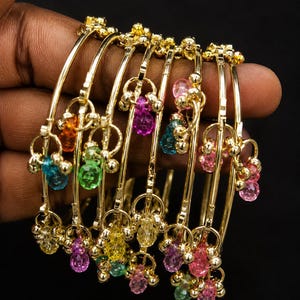 May include: A collection of gold-colored bangles, each adorned with colorful, faceted beads in shades of orange, green, blue, pink, and purple. The bangles are held together, showcasing their intricate design and vibrant gemstones. The jewelry is set against a dark background.