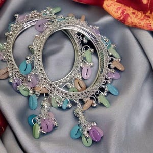 Op de afbeelding: Twee zilveren armbanden met kleurrijke, bungelende kralen in tinten blauw, groen, roze en paars. De armbanden hebben ingewikkelde details en liggen op een grijze satijnen stof.