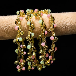 May include: Gold-toned bracelets with a delicate design. Each bracelet features a series of small, round gold beads and is adorned with pink and light green beads. The bracelets are displayed against a dark background.