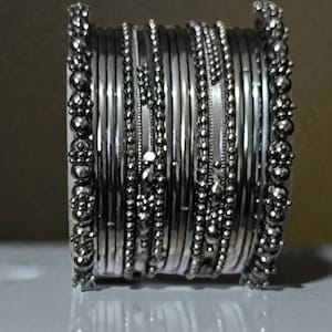 May include: A stack of silver bangles. The bangles are a mix of smooth, beaded, and textured designs. The ends of the stack are decorated with a series of small, round, beaded accents. The jewelry is displayed on a white surface.