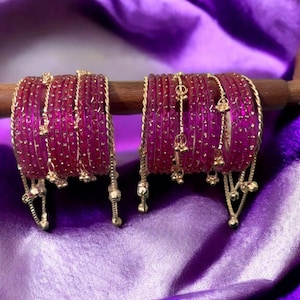 Op de afbeelding: Twee sets magenta armbanden met goudkleurige accenten en bungelende bedels. De armbanden zijn gestapeld en gerangschikt op een houten staaf, tegen een achtergrond van een paarse satijnen stof.