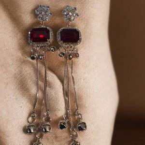 Puede incluir: Un par de pendientes colgantes plateados. Cada pendiente presenta una piedra preciosa rectangular de color rojo oscuro rodeada de piedras claras más pequeñas. Cadenas plateadas y pequeños dijes cuadrados cuelgan debajo.