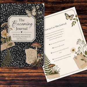 May include: A journal with a black cover featuring the title "The Becoming Journal" in elegant script. The cover is decorated with floral and butterfly illustrations. The open page displays instructions for use, also adorned with botanical elements.