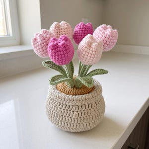 Peut inclure: Un arrangement de tulipes au crochet composé de six fleurs dans des tons de rose clair et rose foncé. Chaque tulipe possède des feuilles et des tiges vertes en crochet, le tout placé dans un pot rond en crochet beige avec une surface supérieure texturée de couleur fauve.