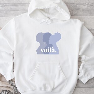 May include: White hooded sweatshirt with a front pocket and drawstring. The front features a design of three women's silhouettes in shades of purple, with the word "voil&agrave;" in a matching purple font.