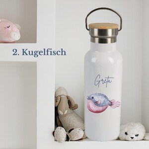 May include: A white metal water bottle with a bamboo lid and a silver handle. The bottle features a watercolor illustration of a pufferfish and the name Greta printed in blue script. The text 2. Kugelfisch is visible to the left of the bottle.