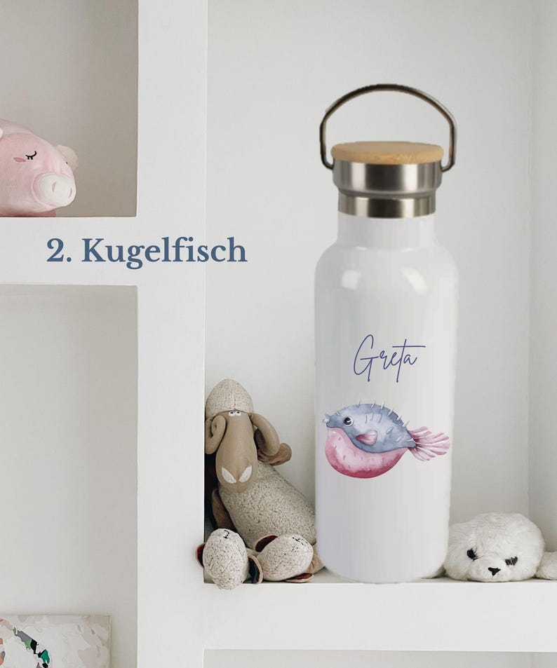 May include: A white metal water bottle with a bamboo lid and a silver handle. The bottle features a watercolor illustration of a pufferfish and the name Greta printed in blue script. The text 2. Kugelfisch is visible to the left of the bottle.