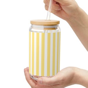 May include: A clear glass tumbler with a cylindrical shape featuring a vertical yellow and white striped pattern. The glass includes a light-colored wooden lid with a central hole and a straight, transparent glass straw inserted through the top.