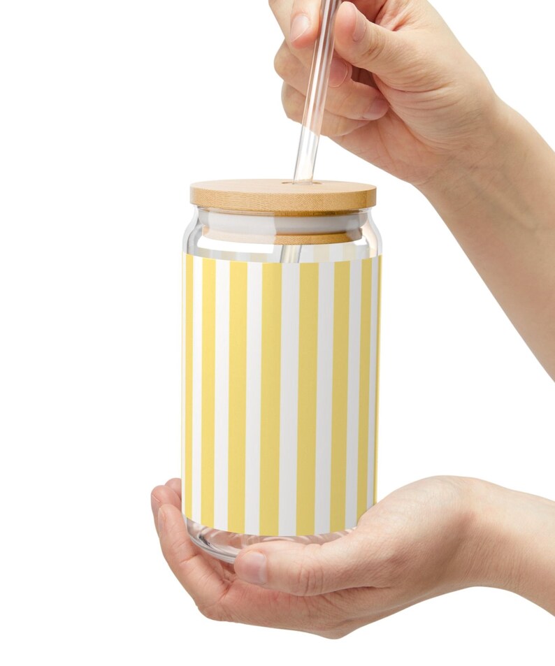 May include: A clear glass tumbler with a cylindrical shape featuring a vertical yellow and white striped pattern. The glass includes a light-colored wooden lid with a central hole and a straight, transparent glass straw inserted through the top.