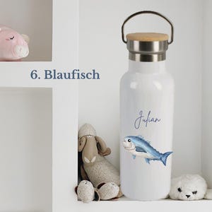 May include: A white water bottle with a silver metal neck and a wooden lid featuring a handle. The bottle displays a blue fish illustration and the name Julian in blue script. The text 6. Blaufisch is visible to the left of the bottle.
