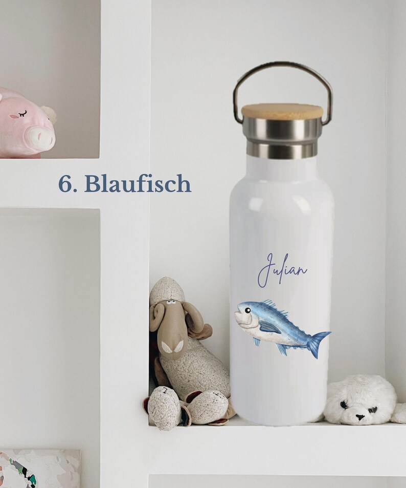 May include: A white water bottle with a silver metal neck and a wooden lid featuring a handle. The bottle displays a blue fish illustration and the name Julian in blue script. The text 6. Blaufisch is visible to the left of the bottle.