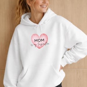 Puede incluir: Sudadera con capucha blanca con bolsillo delantero y cordón. El diseño presenta un corazón rosa con las palabras "MOM is my Inspiration" en escritura negra. La sudadera está hecha de un material suave.