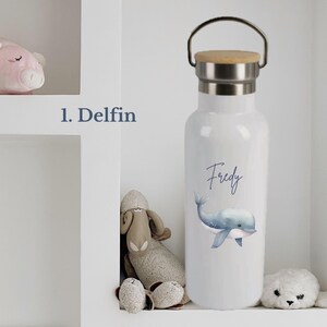 May include: A white metal water bottle with a silver neck and a wooden lid featuring a metal handle. The bottle displays a blue watercolor illustration of a dolphin and the name Fredy in blue script text. The text 1. Delfin is visible nearby.