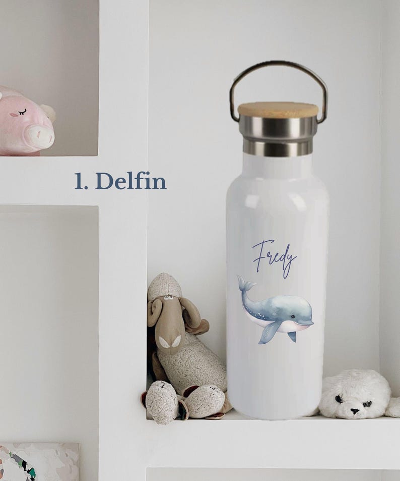 May include: A white metal water bottle with a silver neck and a wooden lid featuring a metal handle. The bottle displays a blue watercolor illustration of a dolphin and the name Fredy in blue script text. The text 1. Delfin is visible nearby.