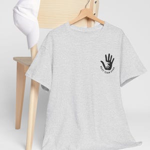 Puede incluir: Una camiseta gris claro con un gr&aacute;fico negro de manos entrelazadas y el texto "BEST TEAM EVER". Una gorra de b&eacute;isbol blanca descansa sobre una silla de madera. La camiseta est&aacute; colgada en una percha de madera.