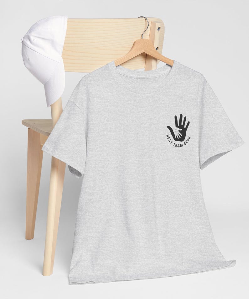 Puede incluir: Una camiseta gris claro con un gr&aacute;fico negro de manos entrelazadas y el texto "BEST TEAM EVER". Una gorra de b&eacute;isbol blanca descansa sobre una silla de madera. La camiseta est&aacute; colgada en una percha de madera.