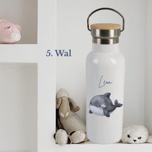 May include: A white water bottle with a silver metal neck and a light brown wooden lid featuring a metal handle. The bottle displays a graphic of a gray and white whale with the name Leon written in blue script above the whale illustration.