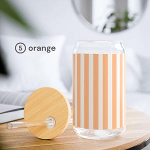 May include: A clear glass tumbler with a vertical orange and white striped pattern, accompanied by a round bamboo lid featuring a central hole and a straight clear glass straw. The text 5 orange is printed to the left of the glass tumbler.