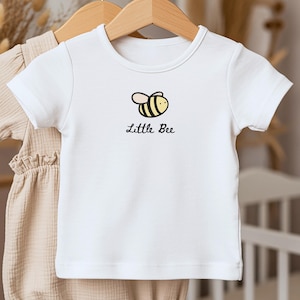 May include: White baby t-shirt with a round neck and short sleeves. The shirt features a cartoon bee graphic in black and yellow, with the words "Little Bee" in a handwritten font below the bee.