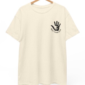Puede incluir: Camiseta color crema con un gr&aacute;fico negro de dos manos, una dentro de la otra, y el texto "BEST TEAM EVER". La camiseta est&aacute; colgada en una percha de madera.