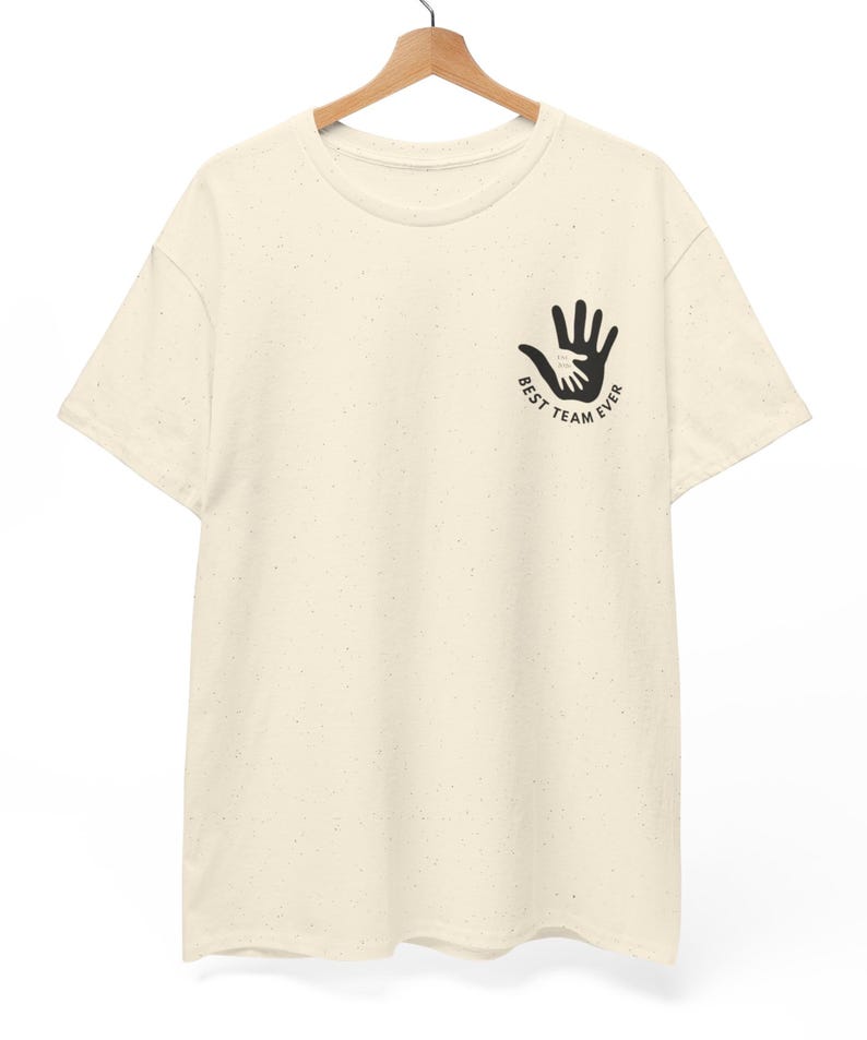 Puede incluir: Camiseta color crema con un gr&aacute;fico negro de dos manos, una dentro de la otra, y el texto "BEST TEAM EVER". La camiseta est&aacute; colgada en una percha de madera.