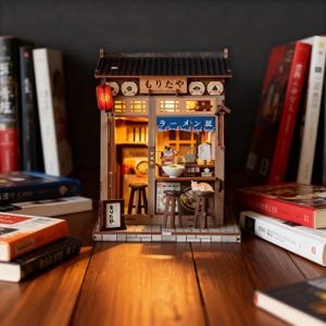 Könnte beinhalten: Miniatur-Modell eines Ramen-Restaurants aus Holz mit detaillierter Fassade, roter Papierlaterne, beleuchtetem Innenraum und japanischem Schild. Das Modell umfasst kleine Hocker, eine Katzenfigur und ist von Büchern umgeben.