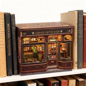 Op de afbeelding: Een gedetailleerd miniatuur Corner Coffee Shop model met bruine gevel en de naam van de winkel boven de ingang. Het interieur is verlicht en toont meubels en decoraties. Het model staat op een witte plank, omringd door boeken.