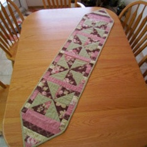 Puede incluir: Un camino de mesa acolchado verde y rosa con un patrón floral. El camino de mesa tiene un extremo puntiagudo y mide aproximadamente 76 cm de largo.