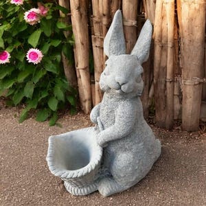May include: A gray bunny statue holding a basket planter. The rabbit has long ears and is seated on a paved surface. Pink flowers and green foliage are visible in the background, with a wooden fence.