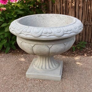 May include: A gray, ornate concrete planter with a wide, scalloped rim and a pedestal base. The planter is round with a bulbous body and sits on a square base. The background includes pink flowers and a wooden fence.