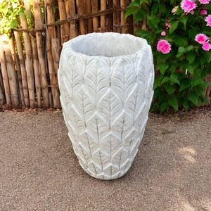 May include: A large, white, outdoor planter with a leaf pattern carved into the surface. The planter is oval-shaped and sits on a paved surface. Pink flowers and a wooden fence are in the background.