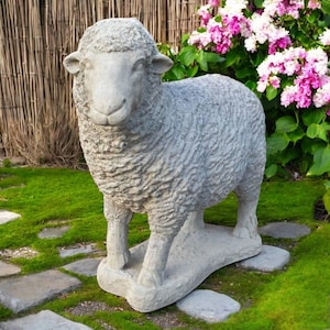 May include: A gray stone sheep statue standing on a stone base. The sheep has a textured woolly coat and is positioned on a bed of green grass and stone pavers. Pink and white flowers are in the background.