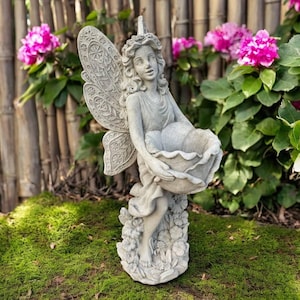 May include: A gray fairy statue with detailed wings and a floral-shaped bowl. The fairy is holding the bowl, standing on a base with floral accents. The statue is outdoors, surrounded by green grass and pink flowers.