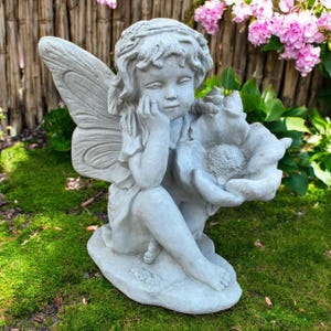 May include: A gray fairy statue with detailed wings and a floral accent. The fairy is seated, resting her chin on her hand, and holding a large flower. The statue is made of a stone-like material and is placed on a bed of green moss.