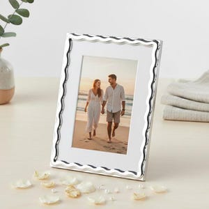 May include: A silver-coloured photo frame with a wavy edge, displaying a picture of a couple on a beach. The frame is angled on a light surface, with scattered petals and a small vase with greenery.