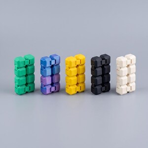 Puede incluir: Cinco juguetes fidget rectangulares dispuestos en fila, cada uno compuesto por m&uacute;ltiples segmentos interconectados en forma de cubo. Los colores de izquierda a derecha son verde s&oacute;lido, un degradado p&uacute;rpura y azul, amarillo s&oacute;lido, negro s&oacute;lido y blanco hueso.