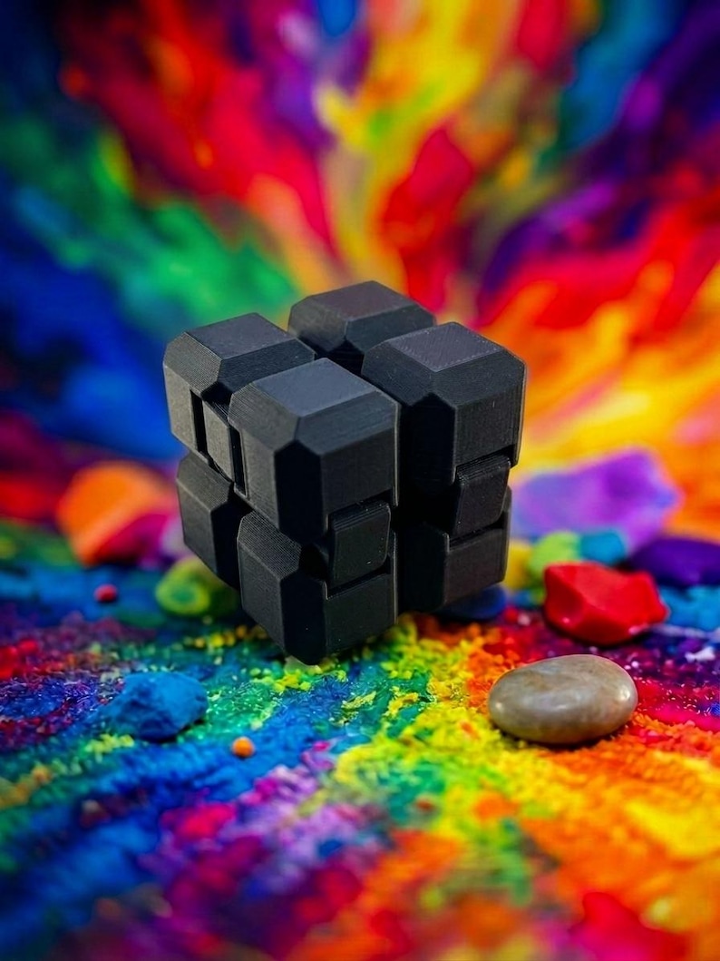 Puede incluir: Un cubo infinito negro mate, juguete antiestr&eacute;s compuesto por ocho cubos peque&ntilde;os conectados con bordes biselados. El objeto presenta un dise&ntilde;o modular que permite plegarlo y desplegarlo, presentado sobre un fondo vibrante y multicolor.