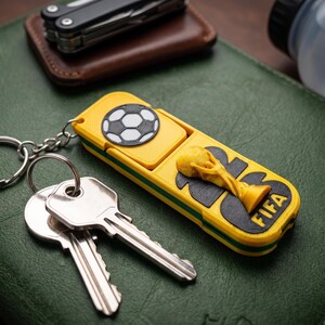 May include: A yellow rectangular keychain featuring a black and white soccer ball graphic, a raised yellow FIFA World Cup trophy emblem, and the text FIFA in black. The keychain is attached to a silver metal ring holding two silver keys.