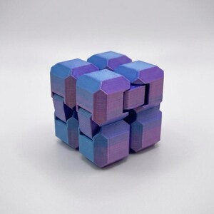 Puede incluir: Un cubo fidget articulado construido a partir de bloques interconectados. La superficie presenta un degradado de color que cambia entre tonos azules y morados. Cada bloque individual tiene bordes biselados y una textura de impresi&oacute;n 3D.