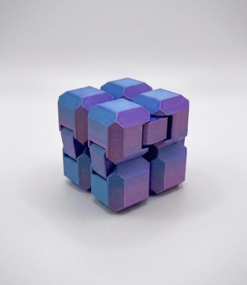 Puede incluir: Un cubo fidget articulado construido a partir de bloques interconectados. La superficie presenta un degradado de color que cambia entre tonos azules y morados. Cada bloque individual tiene bordes biselados y una textura de impresi&oacute;n 3D.