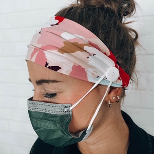May include: A pink and white floral headband with a white button and elastic strap for holding a face mask in place.