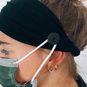 May include: A black headband with a button and white elastic straps. The headband is worn by a person with a green face mask.