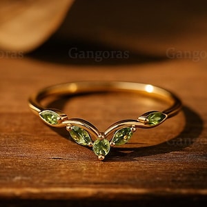 Puede incluir: Un anillo dorado con diseño de hojas. El anillo presenta cinco piedras preciosas ovaladas de color verde claro engastadas en una delicada banda curva. El anillo se muestra sobre una superficie de madera.