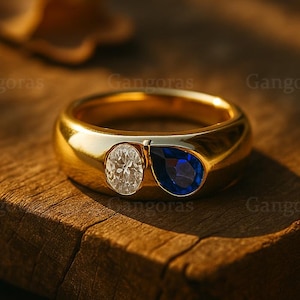 May include: A gold ring featuring an oval white gemstone and a teardrop-shaped blue gemstone. The ring is displayed on a wooden surface, with light reflecting off the polished gold.